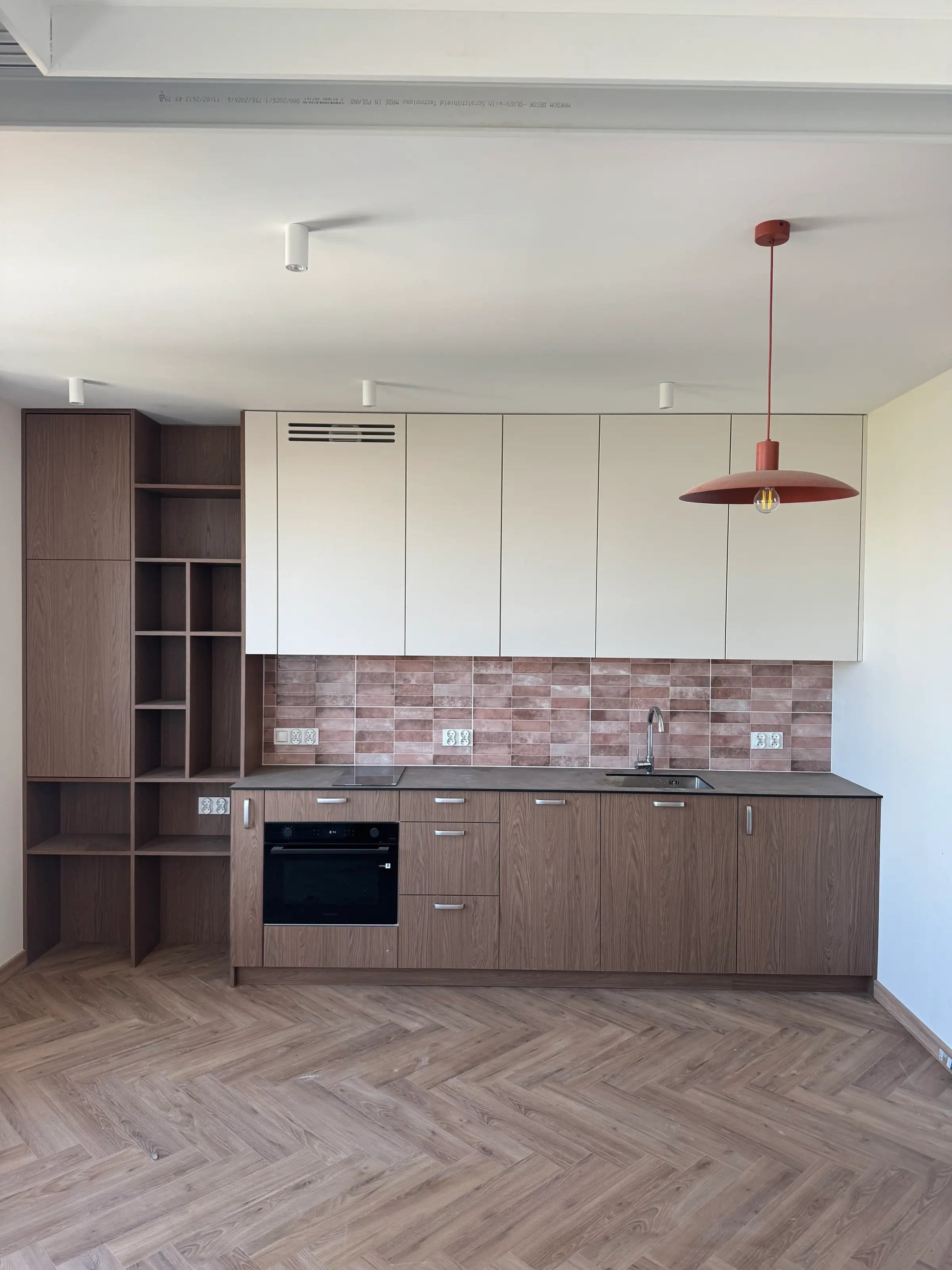Kuchnia z zabudową w kolorze drewna i kremowymi szafkami górnymi, płytki w odcieniach cegły, podłoga w jodełkę i czerwona lampa wisząca.